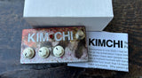 A vintage-style Kimchi fuzz guitar pedal with a distressed, rusted metal body, three cream knobs, and a gold footswitch, shown next to its white box and a printed Kimchi magazine insert on a dark wooden floor