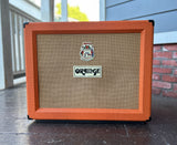 An Orange AD 30 combo amplifier cabinet with a bright orange frame and tan wire mesh grille, photographed outdoors on a porch with a brick wall and blue-gray siding in the background.