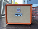 An Orange guitar amplifier cabinet with a bright orange frame and beige woven grille cloth, photographed outdoors on a porch with a brick wall and railing visible in the background.