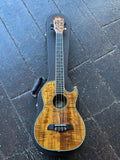 A four-string Baritone Oscar Schmidt  ukulele with a natural flamed Koa wood finish and black hardware, resting on top of its black hard case on a dark wooden plank floor.