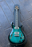 A teal and green PRS electric guitar hollow-body, ebony fretboard with bird inlays with a black hard case, photographed from above on a dark weathered wooden plank surface.