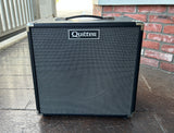 Black Quilter guitar amplifier on a concrete surface with a brick wall in the background