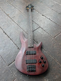 A four-string Schecter  bass guitar with a dark reddish-brown. wood finish,  black hardware, rosewood fretboard wood finish, resting on a dark wooden plank floor.