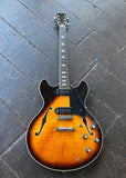 Sire arch-top guitar sunburst finish, laid flat on a dark wooden plank floor. The guitar features a warm orange-to-brown gradient body, a black pickguard, chrome hardware, and a dark fretboard.