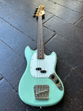 A mint surf green Squier Mustang electric bass guitar with a white pickguard and gold-toned headstock, photographed from above on a dark wooden plank floor.