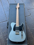 Surh  light blue electric guitar with a black pickguard and maple neck, resting inside a black hard case on a dark wooden deck.