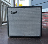 A Supro Black Majick amplifier, guitar amplifier cabinet with a black frame and a gray and white striped grille, photographed outdoors on a porch.