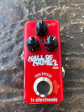 A RED TC Electronic Hall of Fame 2 Mini guitar effects pedal with a bright red body, black knobs, and white lettering, photographed from directly above on a textured dark surface.