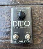 A TC Helicon Ditto Mic Looper guitar effects pedal with a black metal casing, black loop level knob, and silver footswitches, resting on a dark wooden surface.