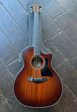 A Taylor model 324ce acoustic-electric guitar with a cutaway body and a brown sunburst finish, resting on a dark wooden plank floor next to its textured gray hard case.