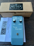 A Sola Sound Tone Bender guitar pedal in a teal blue color, displayed with its original cardboard box, paper insert, and instruction booklet on a dark wooden surface. The box features a black Macarr's brand label.