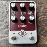 A Universal Audio Ruby Top Boost Amplifier guitar pedal, featuring a maroon red faceplate and a silver aluminum body, resting on a textured brown surface.
