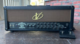 A black Voodoo amplifier head with a gold logo and front control knobs, photographed outdoors on a stone surface with a brick wall and railing in the background.