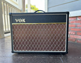 A Vox AC15 guitar amplifier with a black front grille and a brown diamond-pattern speaker grille, photographed outdoors on a porch.
