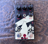 A Walrus black and white Fuzz Face style guitar effects pedal labeled 'MONUMENT' with black knobs, silver footswitches, and red text, photographed from above on a textured brown surface