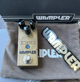 Wampler Tumnus guitar effects pedal in a gold-colored metal casing with black knobs and a silver footswitch, displayed on a gray cloth next to a Wampler sticker and in front of a black Wampler box.