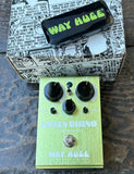 A Way Huge Green Rhino guitar effects pedal in lime green with black knobs, shown in front of its comic-book-patterned box and a black carrying case on a dark stone surface.